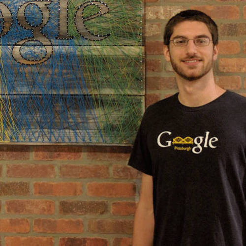 A man wearing glasses and a black 'Google Pittsburgh' t-shirt standing in front of a brick wall with a colorful string art Google logo.